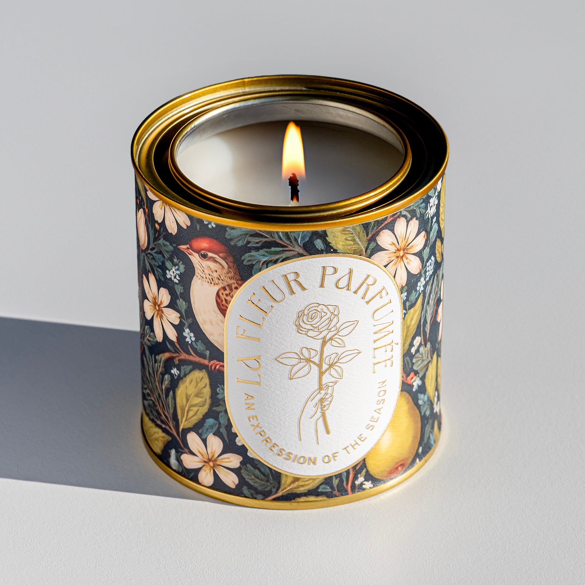 An elegant luxe lit gold-tone candle,  wrapped in artwork of a serene winter woodland scene featuring majestic white-tailed deer congregating on snow-covered terrain, framed by tall evergreens. The soft, neutral tones of the trees complement the snow-covered earth, evoking a calm atmosphere. The composition captures a tranquil winter day, with the wildlife enhancing the sense of natural beauty amidst the crisp, cool landscape.