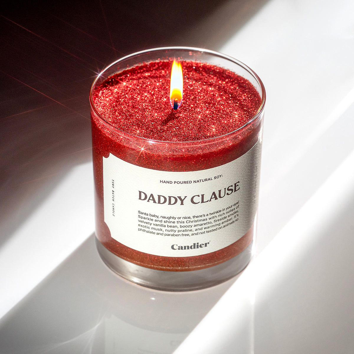 A lit festive Christmas red scented candle with plant based sparkly glitter and a label that reads Daddy Clause by Candier 