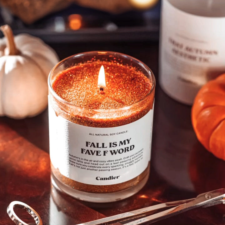 A sparkling autumn colored candle with plant based glitter and a label that reads Fall Is My Fave F Word, in a cozy fall decorated festive setting