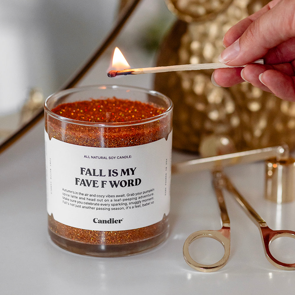 A sparkling glittery fall scented candle with a label that reads Fall Is My Fave F Word. It is being lit with a match and has a gold candle snuffer and trimmer beside it