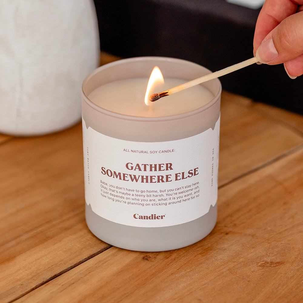 A luxe fall scented candle being lit with a match, with a label that reads Gather Somewhere Else