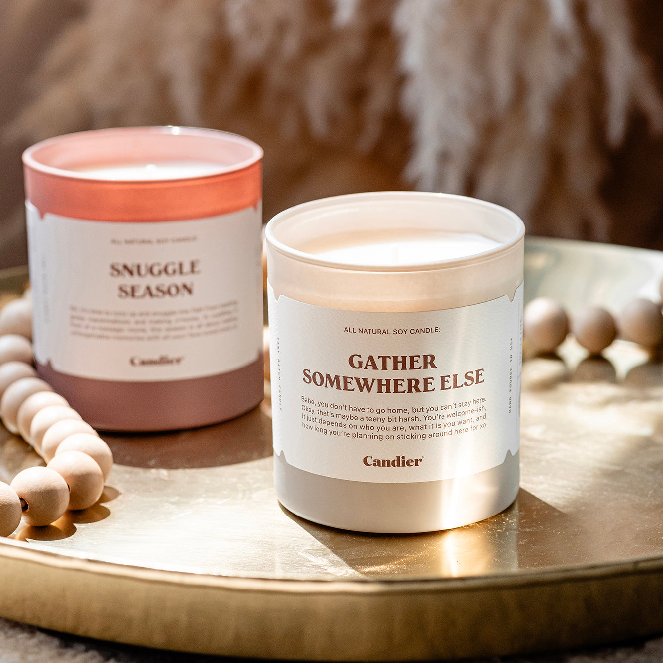 A luxe fall scented candle with a label that reads Gather Somewhere Else, with another candle that reads Snuggle Season, surrounded by elegant fall decor