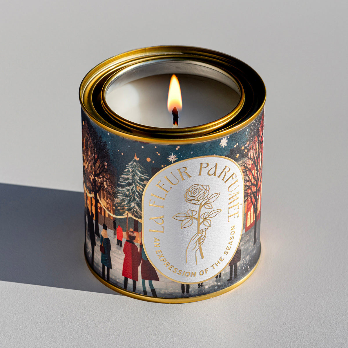 An elegant luxe lit gold-tone candle depicting townsfolk gathering to celebrate Christmas in a charming square under twinkling lights. Snow falls gently as figures in cozy winter wear admire the grand Christmas tree in the town square. The warm glow of shop windows and elegant buildings creates a welcoming ambiance. 