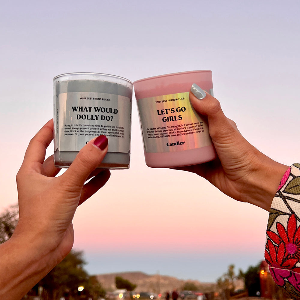 a pink candle held in a woman's hand with a label that reads Let's Go Girls, with a pink sunset 