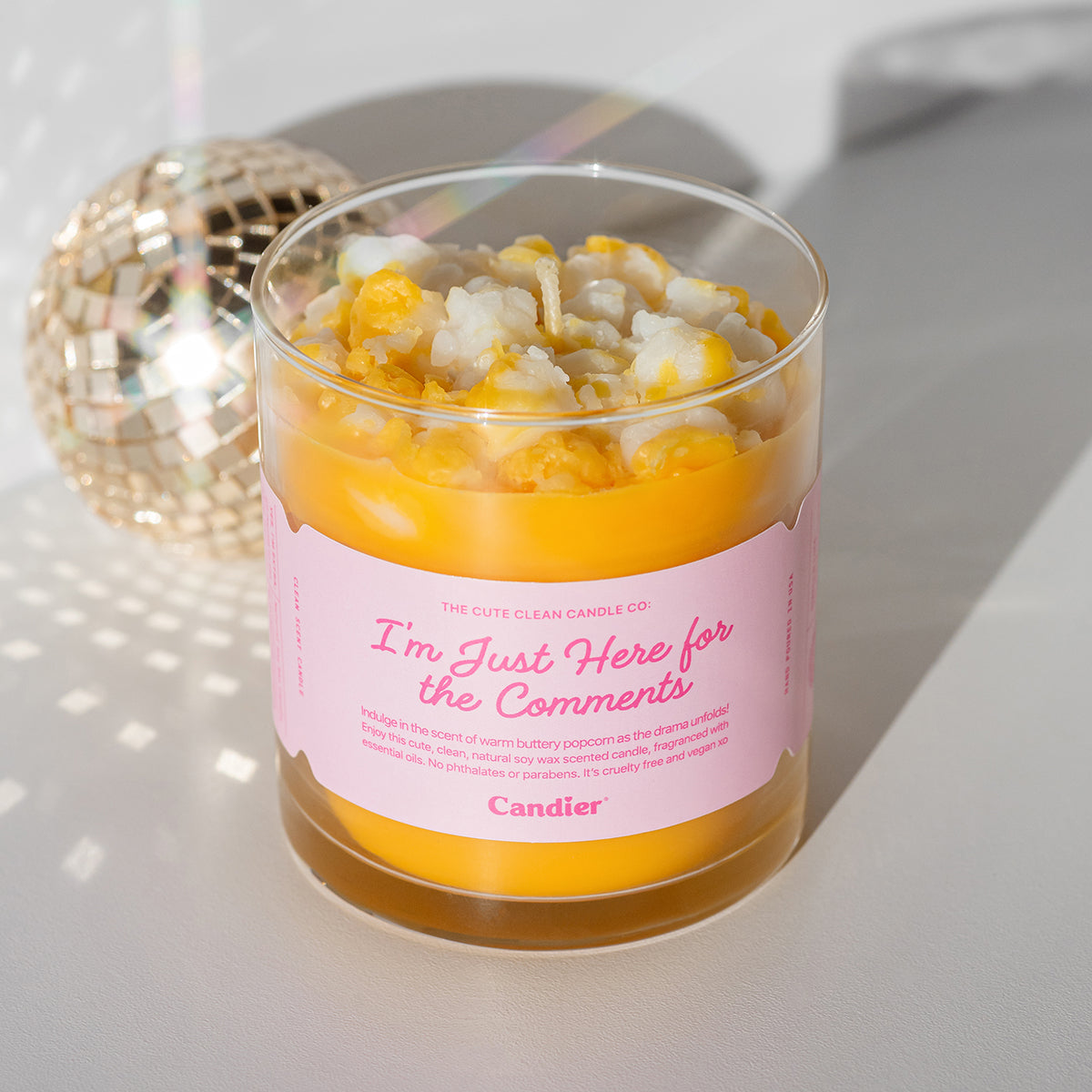 A fun premium scented candle topped with realistic wax popcorn and a label that reads I'm Just Here For The Comments by Candier®, besides a sparkling mirror disco ball