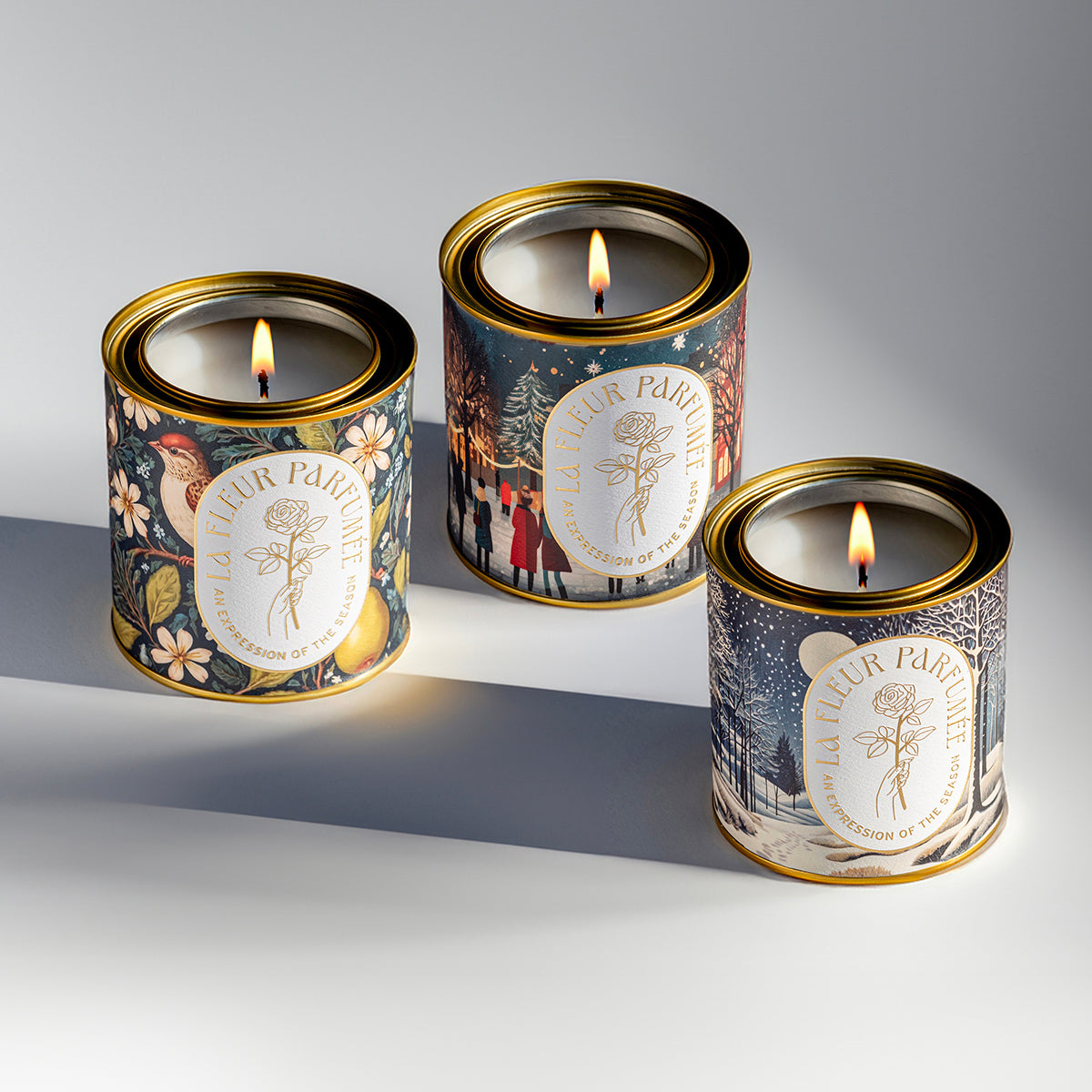 Three Christmas themed luxury candles, one depicting a partridge in a pear tree, one of a cozy Christmas tree lighting ceremony in a festive village scene, and another of a crisp snowy forest