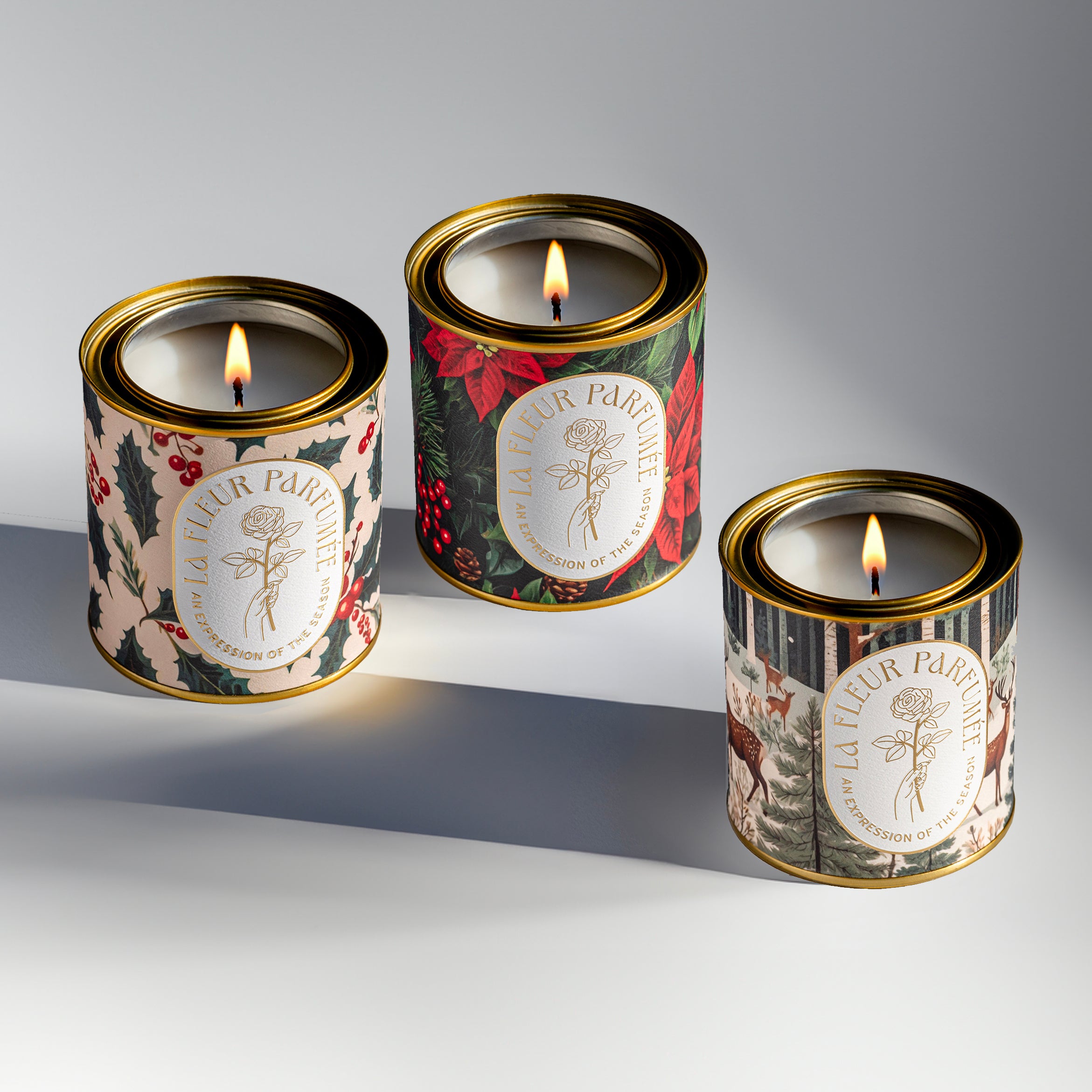 Three luxurious Christmas themed candles with bold holiday inspired artwork, including festive holly, poinsettia, and a snowy woodland scene with whitetail deer