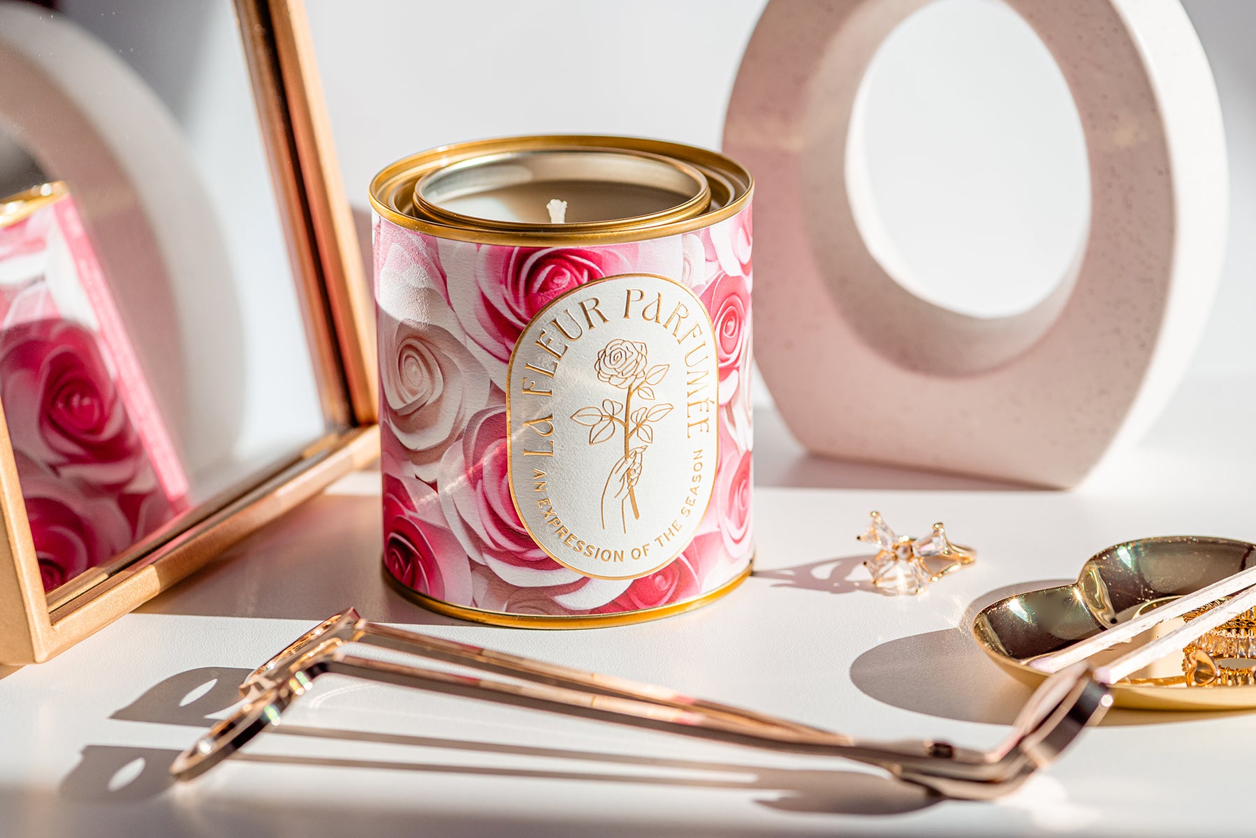 Luxury soy rose candle with elegant gold logo that reads La Fleur Parfumeé An Expression of the Season. The gold tone metal vessel is decorated with artwork of pink and cream roses. The candle sits on a table beside a wick trimmer, heart gold dish and other stylish decor