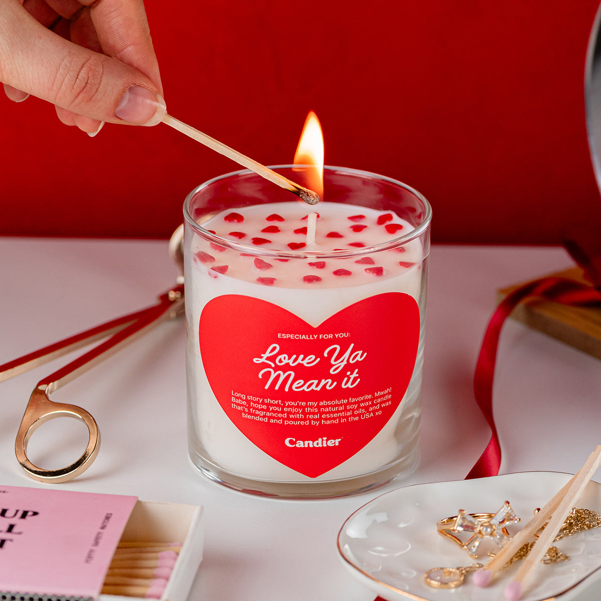 A cute scented candle with heart shaped sprinkles and a heart shaped label that reads Love Ya Mean It by Candier®, being lit with a match