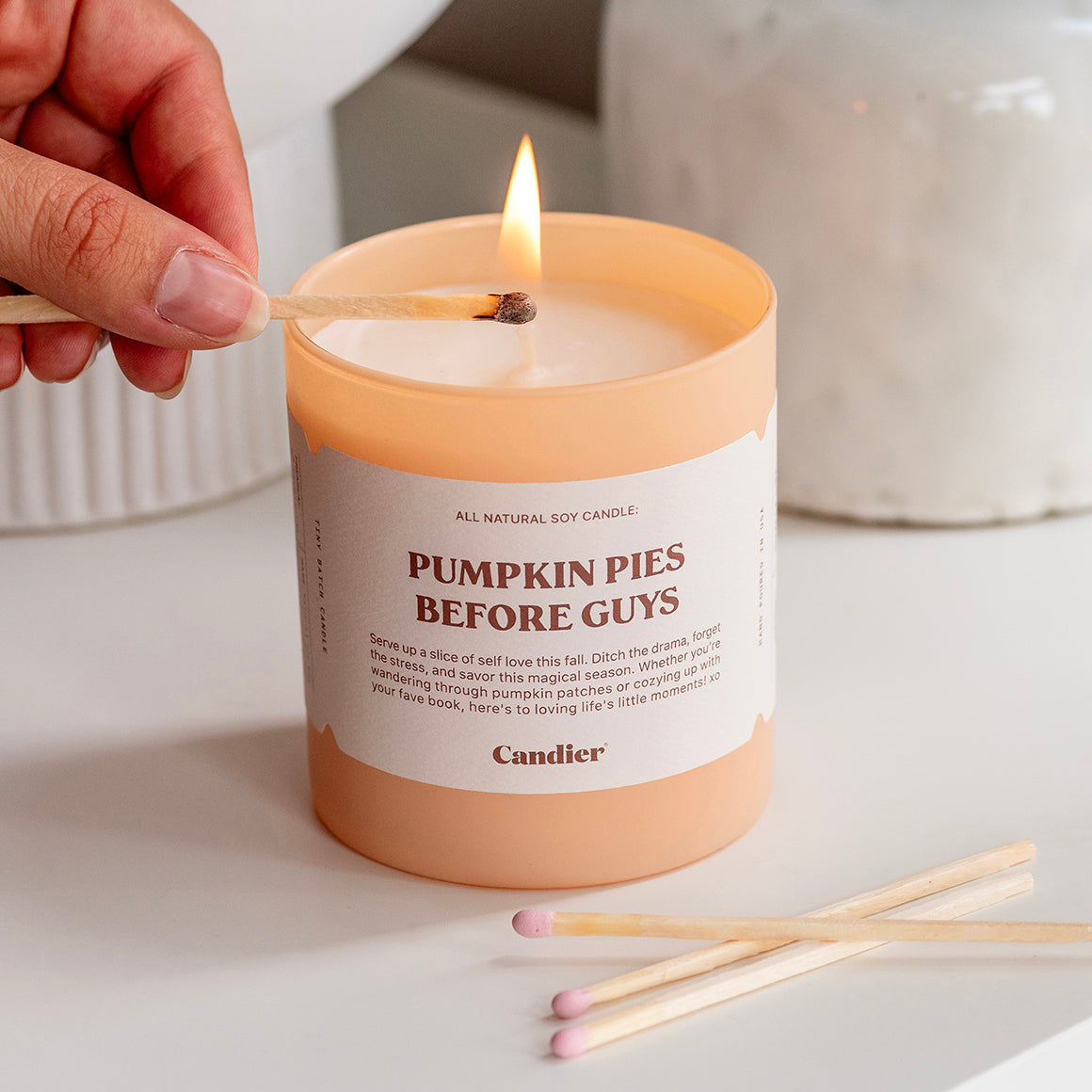 A cute luxury fall scented candle with a label that reads Pumpkin Pies Before Guys, it is being lit by a match