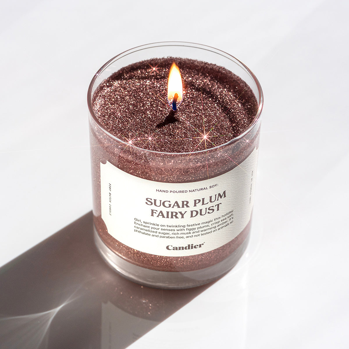 A lit glittery pink Christmas themed scented candle with a label that reads Sugar Plum Fairy Dust by Candier