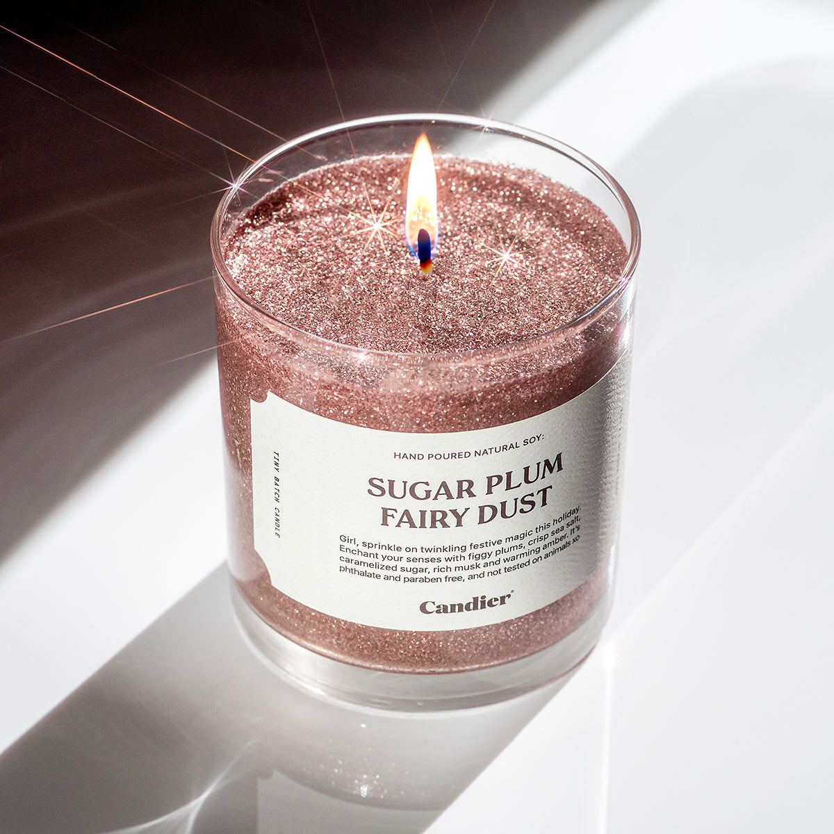 A lit glittery pink Christmas themed scented candle with a label that reads Sugar Plum Fairy Dust by Candier