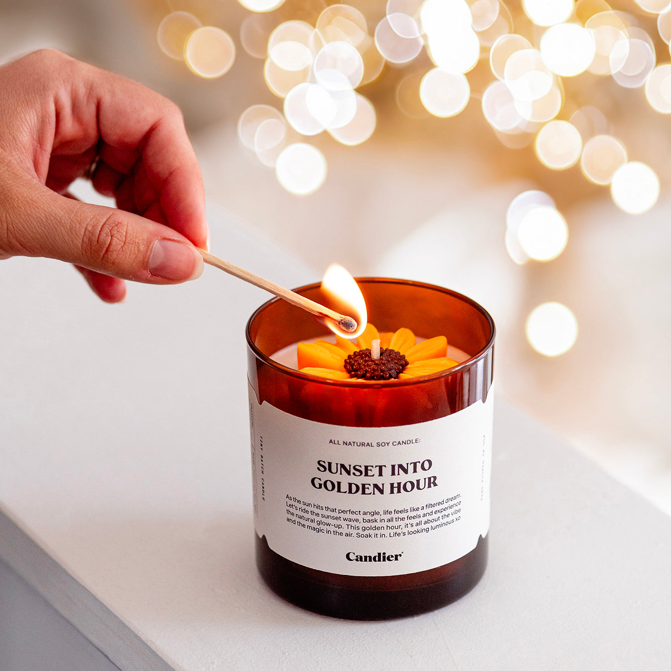 a fall scented candle being lit by a match. It has a ute sunflower detail and a label that redas Sunset Into Golden Hour