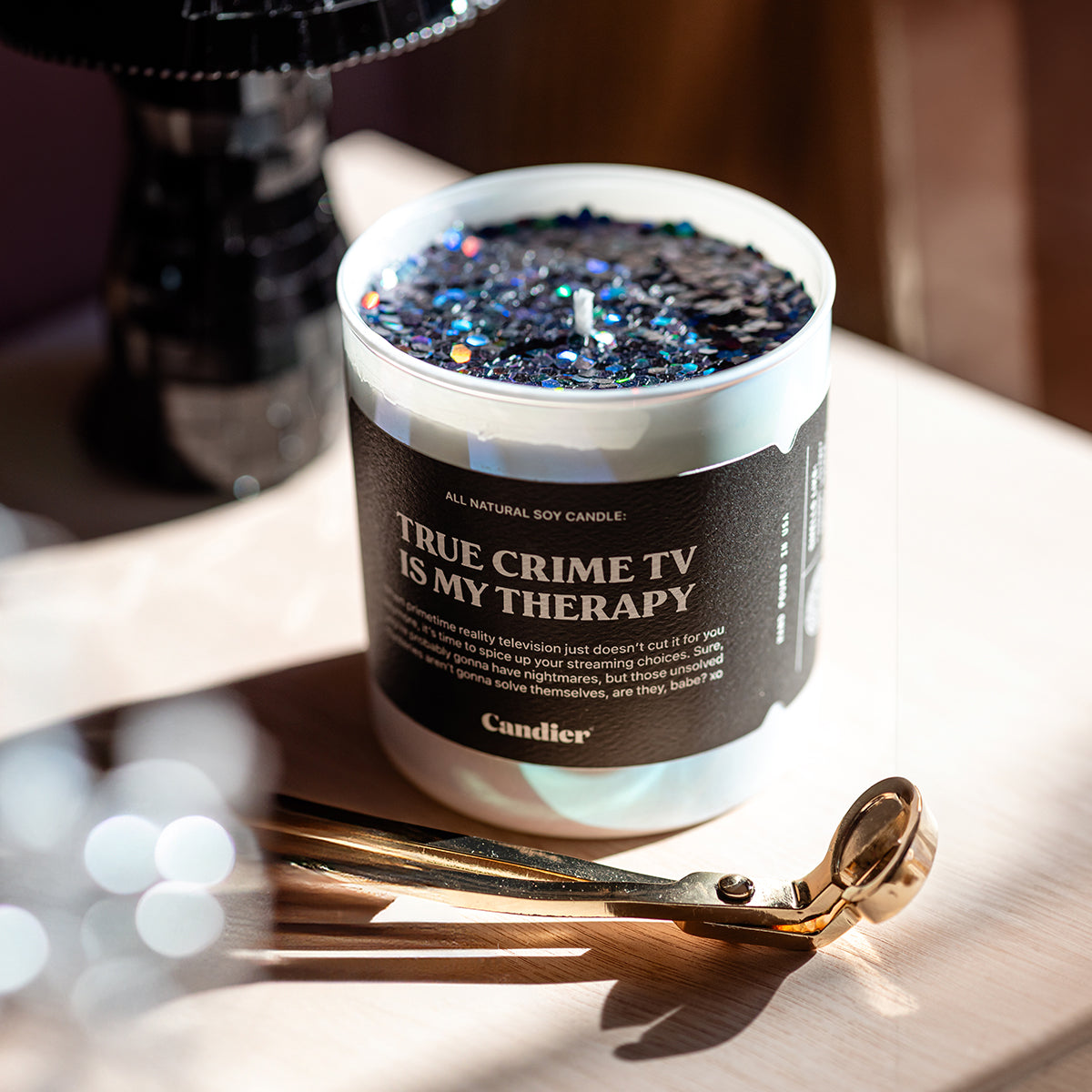 a shiny luxury scented candle, with sparkling plant based candle glitter, and a black label that reads True Crime TV Is My Therapy, in a cozy setting