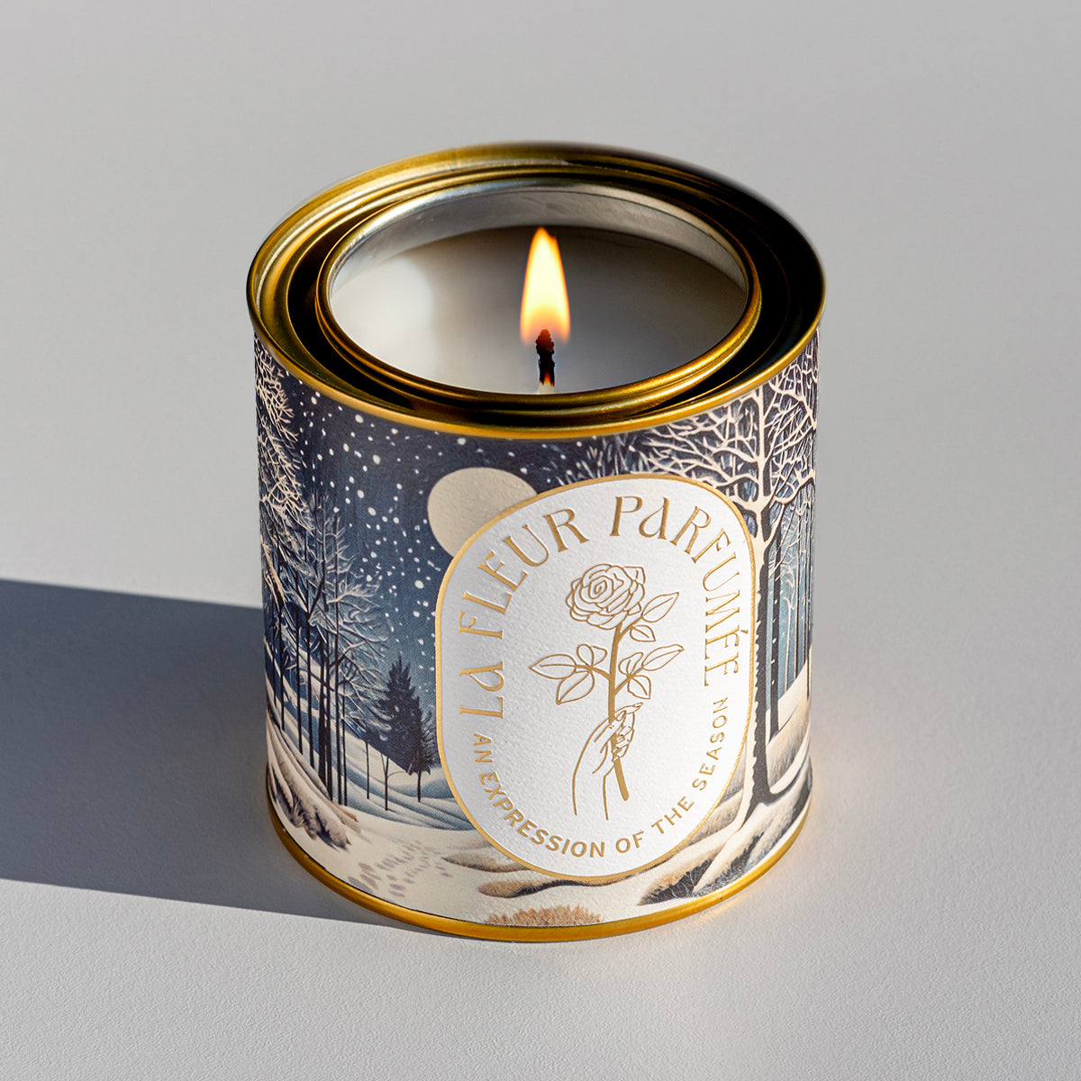 An elegant luxe lit gold-tone candle,  wrapped in artwork of a peaceful winter night scene illuminated by the soft glow of the moon, with snow gently falling over a quiet forest. Bare trees stretch upward, their branches dusted with light snow, while soft snowdrifts blanket the ground. Footprints trail off into the distance, adding a sense of wonder. The composition evokes a serene winter’s eve.