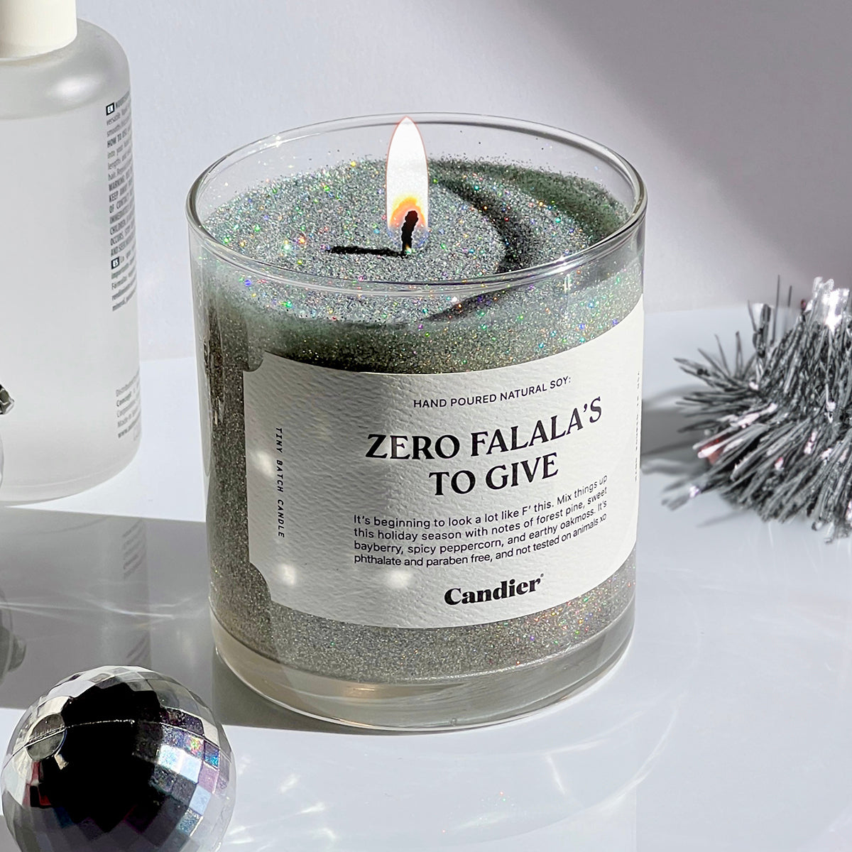 A lit sparkling Christmas themed candle with a label that read Zero Falala's to Give by Candier, topped with plant based eco glitter, with silver Christmas decor