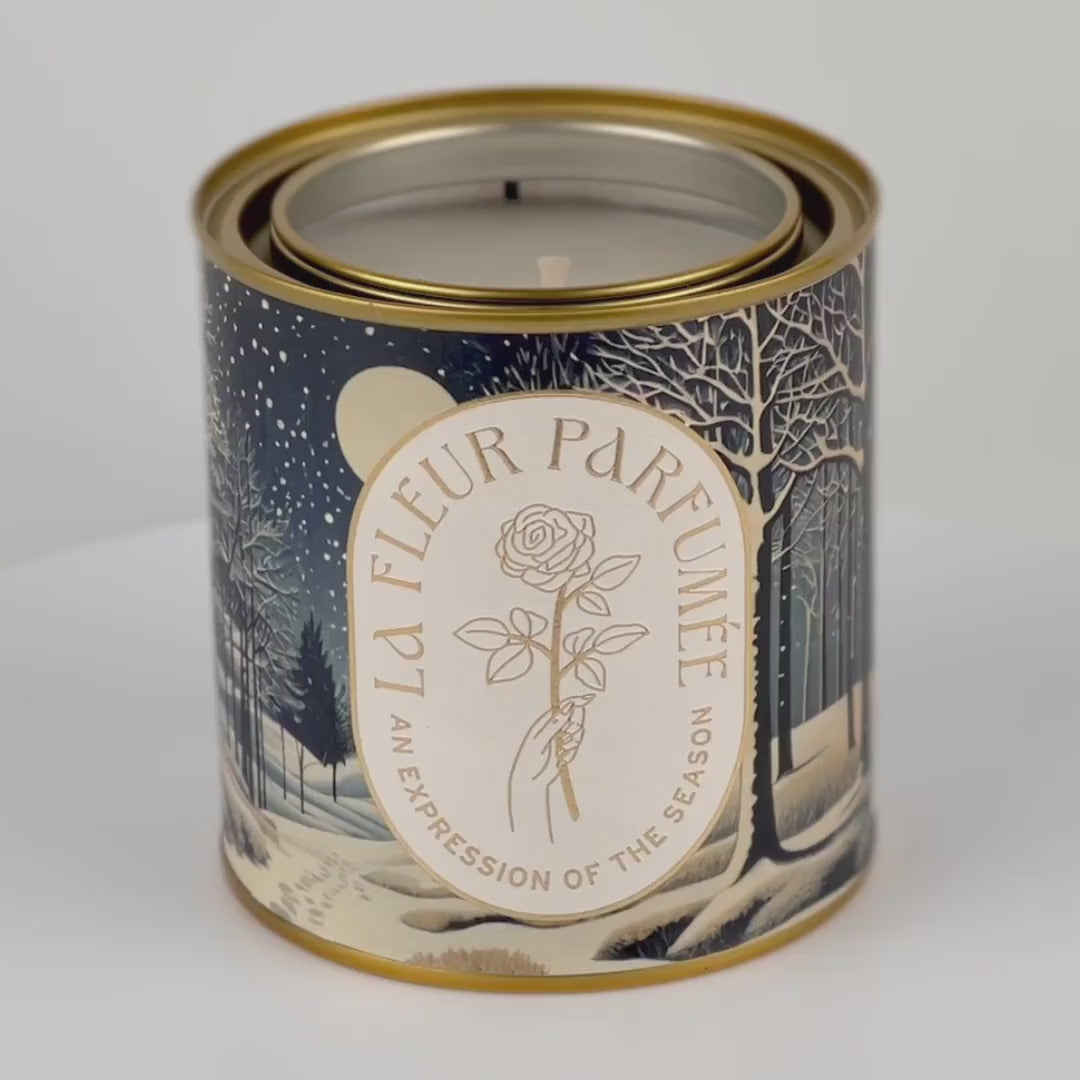 A 360 degree video of a luxury Christmas candle, wrapped in artwork of a peaceful winter night scene illuminated by the soft glow of the moon, with snow gently falling over a quiet forest. Bare trees stretch upward, their branches dusted with light snow, while soft snowdrifts blanket the ground. Footprints trail off into the distance, adding a sense of wonder. The composition evokes a serene winter’s eve.